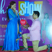 a man is kneeling down in front of a woman holding a red heart shaped balloon in front of a sign that says show