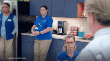 a woman in a blue shirt with the word superstore on it stands in a kitchen with two other women