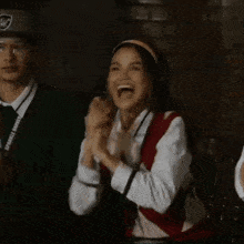 a girl in a school uniform is clapping her hands in the air while standing next to a boy in a baseball cap .