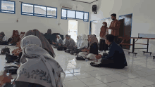 a group of people sit on the floor in a room with a man wearing a mask