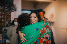 a woman in a green floral shirt is hugging another woman in front of a wall with a sign that says pklta