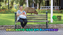 a woman sits on a park bench with a capybara on her lap and the words enquanto isso em blumenau written above her