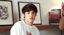 a young man wearing a white shirt and a necklace is sitting in front of a shelf with paintings on it .