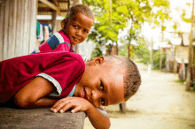 a boy in a red shirt is laying on a wooden bench next to another boy in a striped shirt