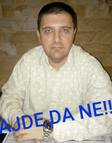 a man wearing a watch is sitting at a table with the words ajde da nei written on the front of his shirt