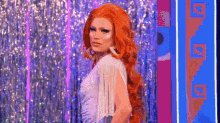 a drag queen with red hair and a white dress is standing in front of a curtain .