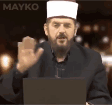 a man with a beard wearing a white hat stands in front of a laptop computer .