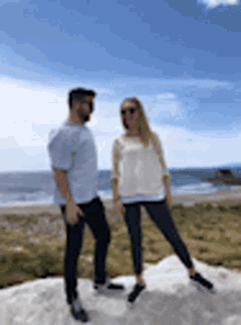 a man and a woman are standing next to each other on top of a rock near the ocean .