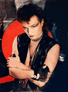 a man in a black leather vest and choker is standing with his arms crossed in front of a red circle .