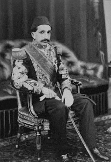 a black and white photo of a man in a suit sitting in a chair with a cane .