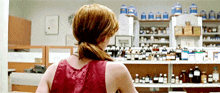 a woman in a red tank top is standing in a pharmacy looking at shelves filled with bottles of medicine .