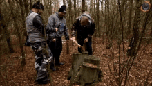 three men are standing around a tree stump in the woods and one of them is wearing a hat that says skull