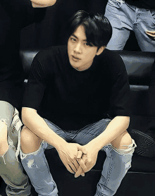 a man in a black shirt and ripped jeans sits on a black couch