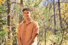 a young man in an orange shirt stands in the woods with his hands behind his back