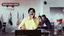 a man with a mustache is sitting at a desk in a classroom with his hand on his face .