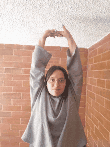 a woman wearing glasses and a grey shirt stretches her arms in front of a brick wall