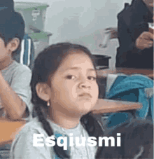 a little girl is sitting at a desk in a classroom with the words esquismi written on the bottom of her face .