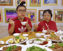 two women are sitting at a table with plates of food and one of them is wearing a red adidas shirt