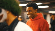 a young man in a red hoodie smiles while talking to another man