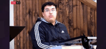 a man wearing glasses and a black adidas jacket is sitting in front of a wooden wall