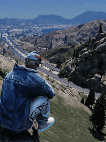 a man in a denim jacket sits on top of a hill overlooking a highway