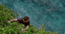 a man is laying on the edge of a cliff overlooking the ocean and a woman is swimming in the water .