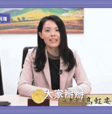 a woman in a pink jacket sits at a desk with a name plate that says ' rainbow ' on it