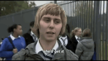 a young man in a school uniform is standing in front of a fence talking to a group of people .