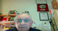 an elderly man wearing glasses is sitting in front of a wall with plaques on it .