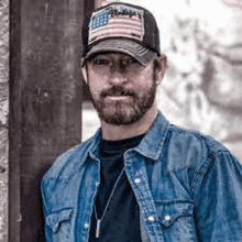 a man with a beard wearing a hat and a denim shirt .