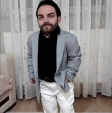 a man with a beard is standing in a living room wearing a jacket and white pants .