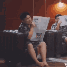 a man sits on a couch reading a newspaper while another man sits next to him reading a book