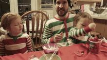 a man in a green and white striped shirt is sitting at a table with two children and a container of strawberry yogurt