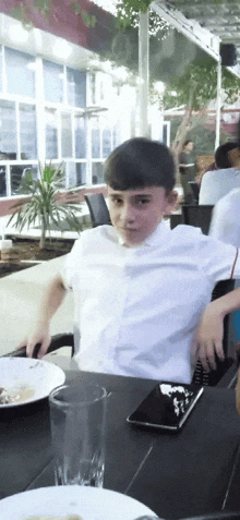 a young boy sits at a table with a cell phone and a glass