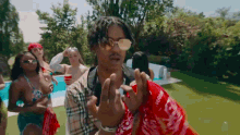 a man wearing sunglasses and a red bandana is standing in front of a pool surrounded by women in bikinis .