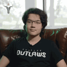 a man wearing a black houston outlaws shirt sits on a couch