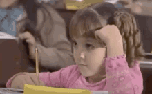 a little girl in a pink shirt is sitting at a desk in a classroom with a pencil in her hand .