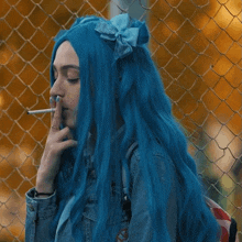 a woman with blue hair is smoking a cigarette in front of a chain link fence