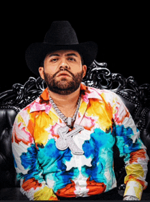 a man wearing a cowboy hat and tie dye shirt is sitting on a chair