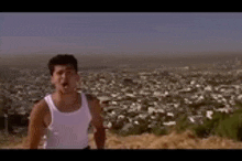 a man in a white tank top is standing in front of a city .