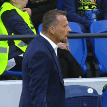 a man in a suit stands in front of a row of blue seats with a yellow vest with the number 3 on it