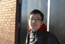 a young man wearing glasses and a red hoodie is standing in front of a brick wall