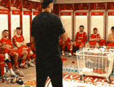 a man in a black shirt stands in front of a group of players in a locker room with the name mac allister on the wall