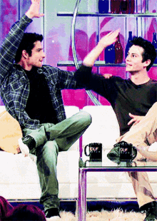 two men are sitting on a couch with their arms in the air and one of them is wearing a plaid shirt