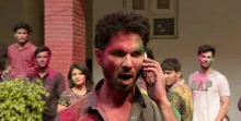 a man with holi paint on his face is talking on a cell phone in front of a crowd of people .