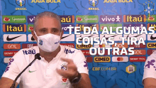 a man wearing a face mask is speaking into a microphone in front of a wall with advertisements for itau