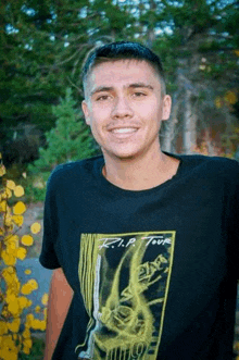 a young man wearing a black t-shirt is smiling and standing in front of a forest .