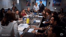 a group of people are sitting around a table in a conference room with a nbc logo on the wall .