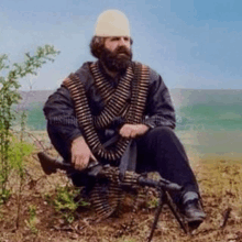 a man with a beard is sitting in the dirt with a gun .