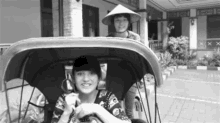 a black and white photo of two women riding in a rickshaw . one of the women is wearing a conical hat .